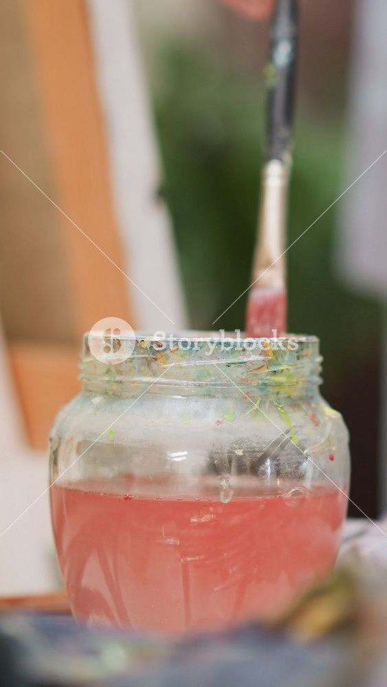 Artist washes brush in water poured into glass jar closeup. Art teacher cleans paintbrush from red pigment at table in shop. Creative man