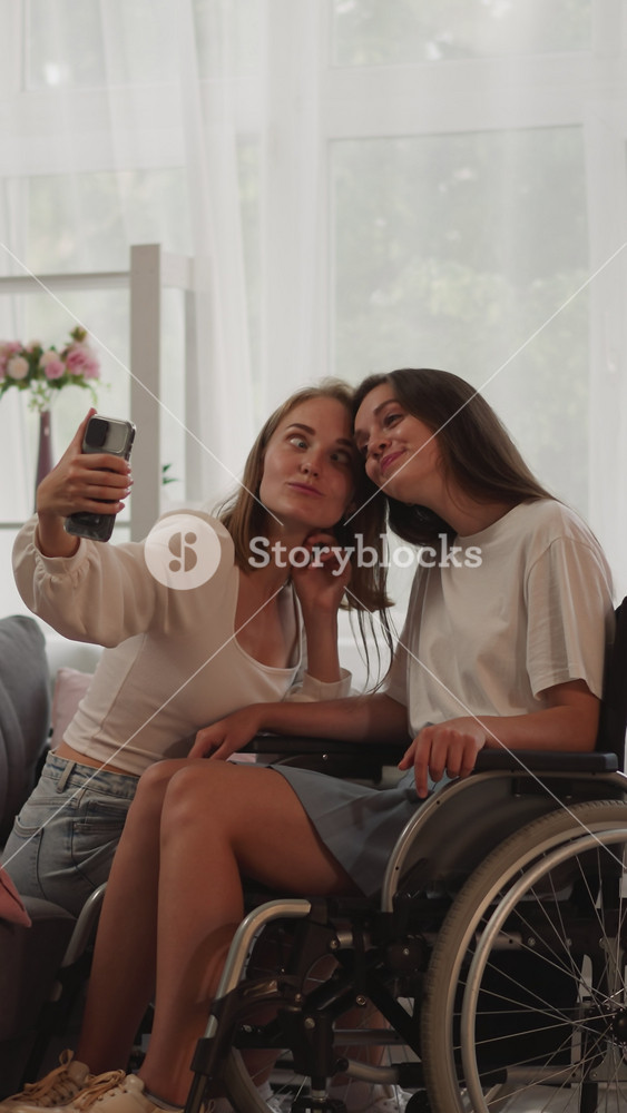 Happy Lesbian couple makes selfie on smartphone sticking out tongue and smiling. Blonde woman enjoys time with partner with disability in