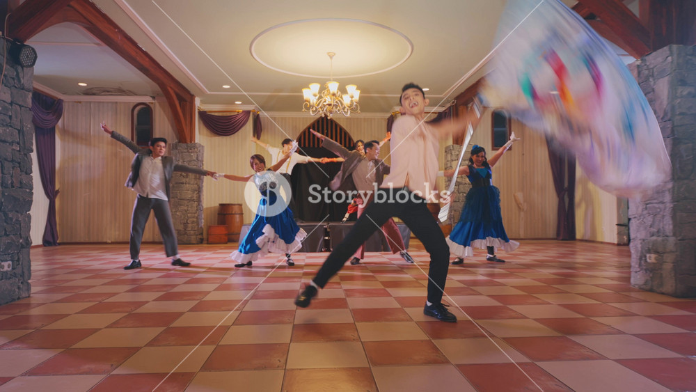 A lively dance scene in a spacious hall with wooden accents. Couples are dancing joyfully, while one person is playfully falling to the floor. Colorful costumes add vibrancy to the atmosphere.