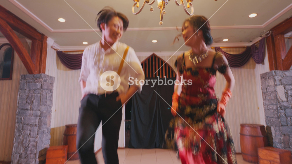 A joyful couple dancing in a beautifully decorated indoor space with a chandelier. The woman wears a colorful dress while the man is in a casual white shirt. The ambiance is warm and inviting.