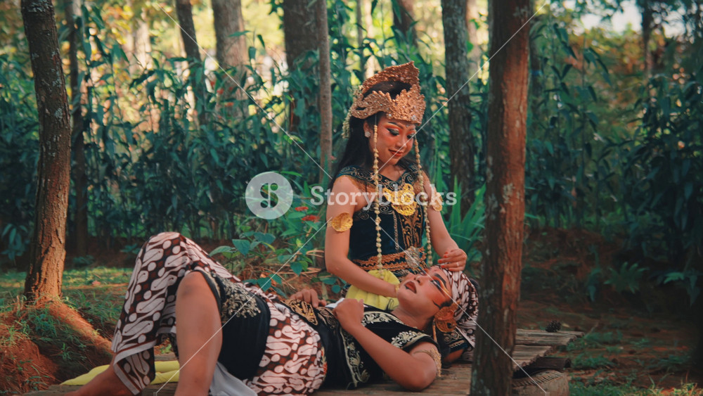A man portraying the mythological character Rama rests on a wooden platform in a forest, his head in the lap of his partner, Sinta. She tenderly cares for him in a beautiful scene of love and folklore.