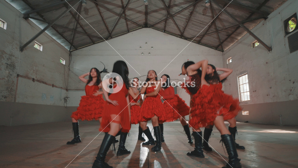 A group of four dancers in red dresses performing in an industrial space. The dancers showcase energetic movements, with a focus on rhythm and style. The background features large fans and a spacious floor.
