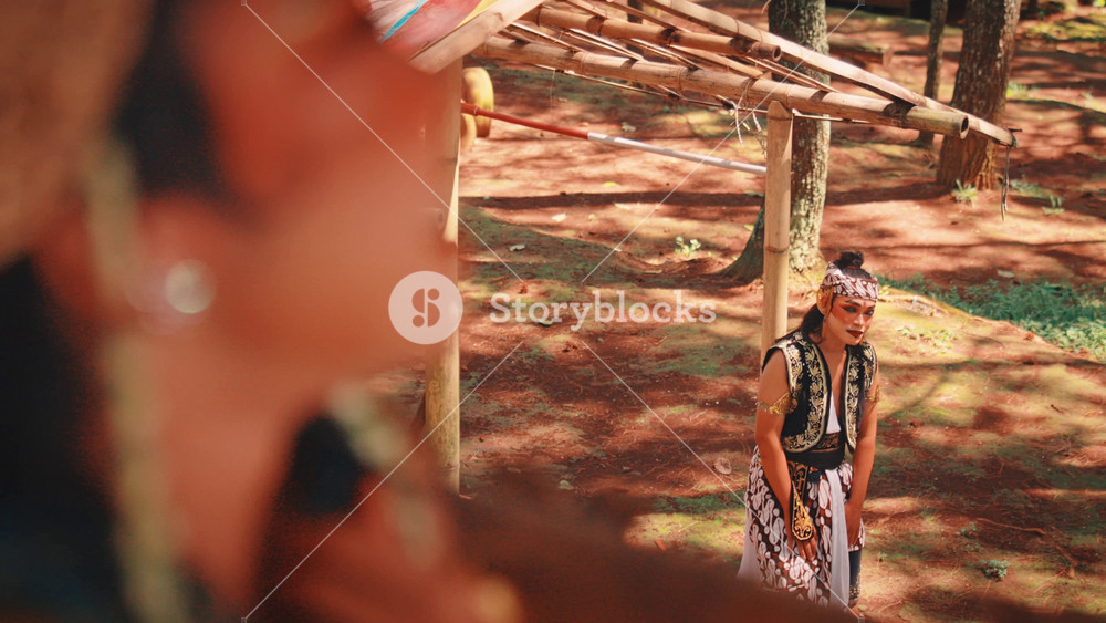 An over-the-shoulder shot showing a female Javanese dancer watching her male counterpart, who stands under a bamboo shelter in the distance. The shot conveys a sense of longing, distance, or observation.