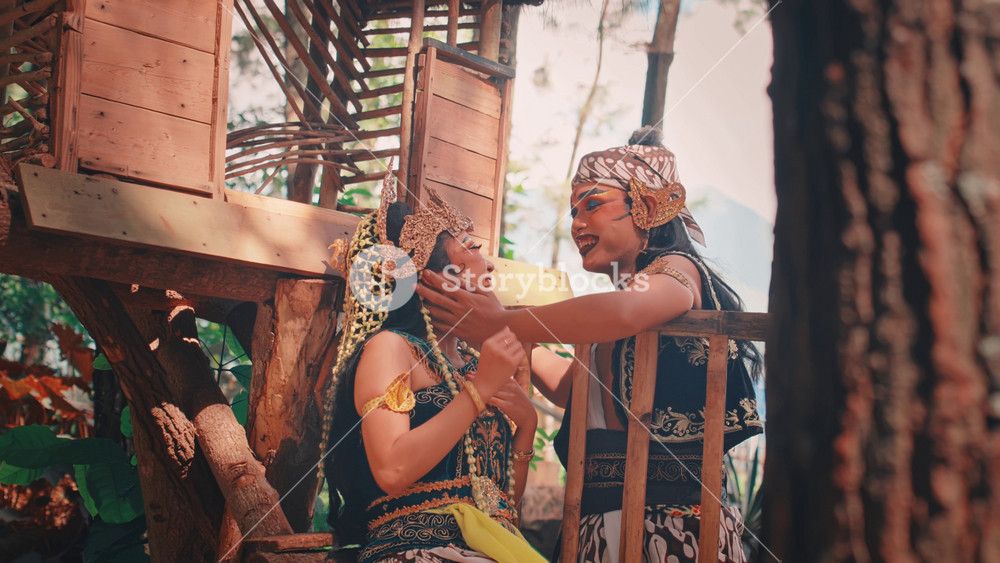 A male and female Javanese dancer in ornate costumes share a playful and intimate moment in a treehouse. They laugh and smile as the man tenderly touches the woman's face.