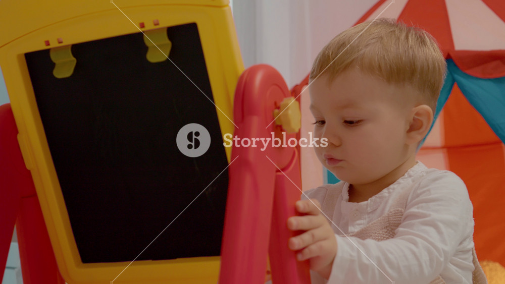 A young child focuses on drawing colorful pictures on a blackboard in a lively playroom. The surroundings are colorful and inviting, sparking creativity.