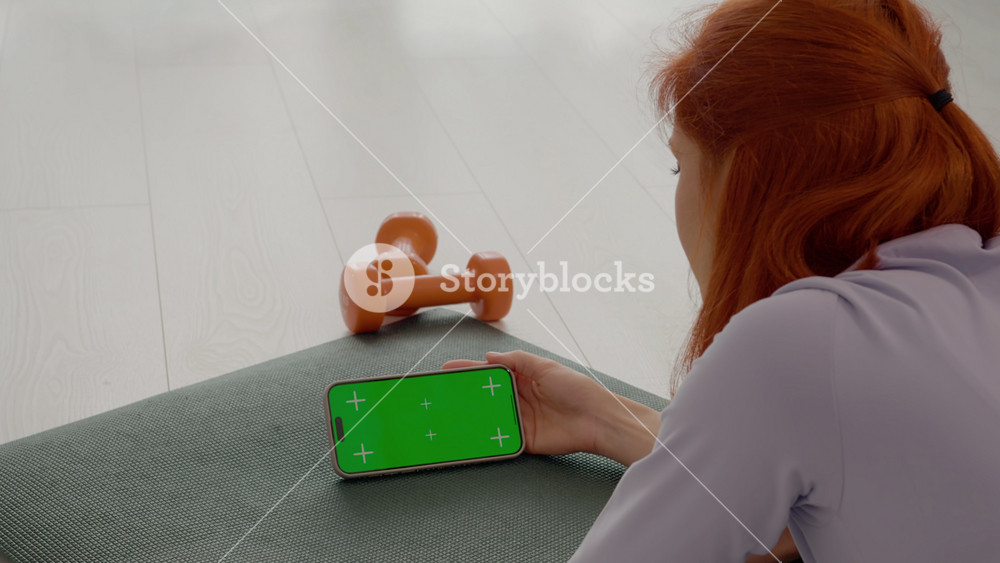 A woman with red hair takes a break on her gym mat, swiping her smartphone with a green screen. She enjoys a moment of relaxation in her living room.