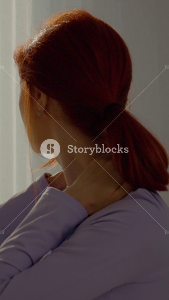 Vertical Screen: A redhead woman sits comfortably in her living room, gently massaging her neck to soothe discomfort from muscle pain caused by strain or dislocation.