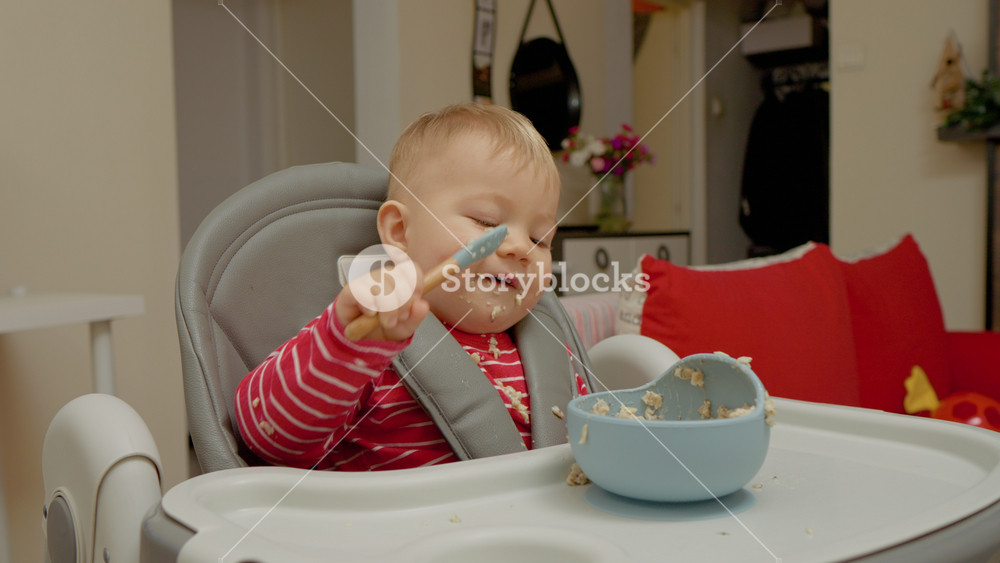 A little boy in a high chair explores the joys of breakfast as he learns to self-feed with a blue bowl of cereal. His playful attitude makes mealtime entertaining for everyone involved.