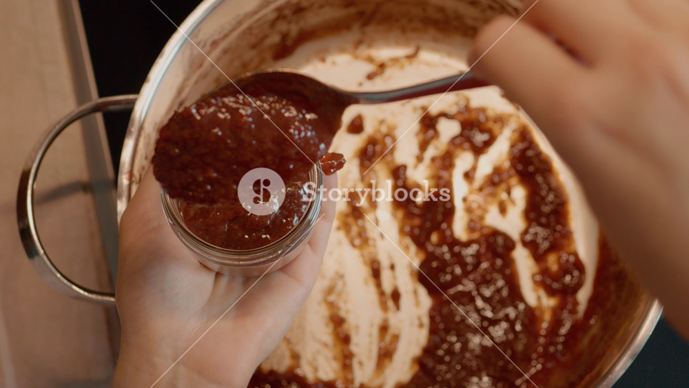 Top view of a woman's hands gently fill glass jars with rich, freshly boiled plum jam, showcasing the art of preserving seasonal fruit for enjoyment throughout the year.