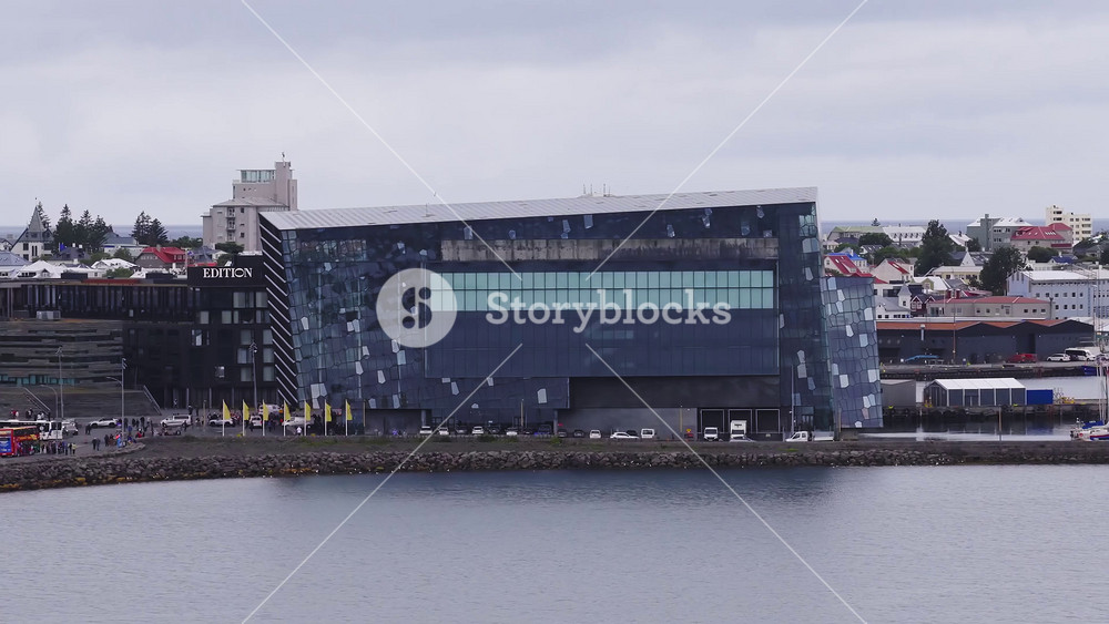 Aerial slow pan of Harpa's angular glass facade, EDITION hotel sign, colorful pitched roof houses, warehouses, and marina in Reykjavik under soft overcast light.