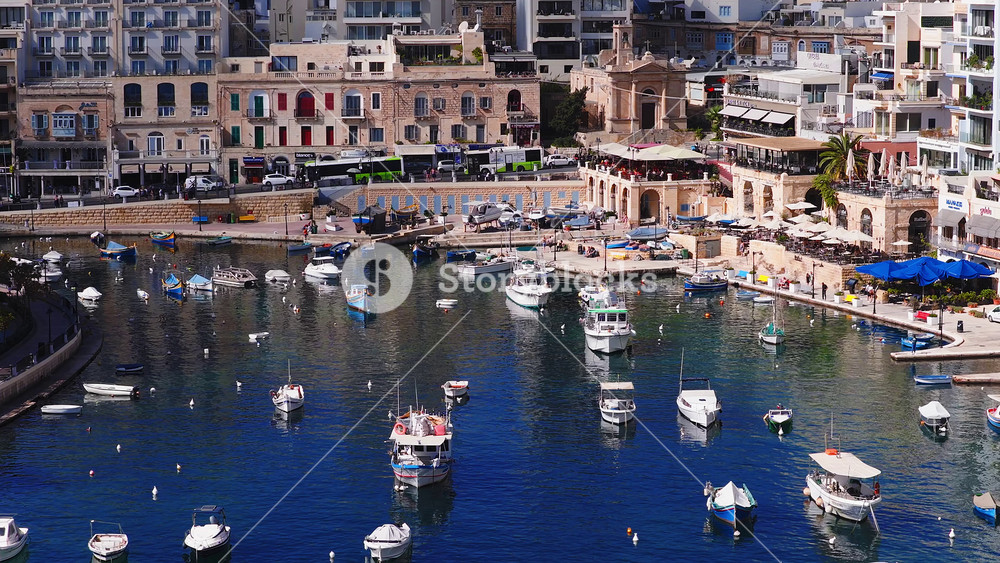 Aerial view of Sliema harbor in Malta shows moored boats, a jet ski, limestone terraces, cafes, a small church facade, and people on the promenade in midday light.