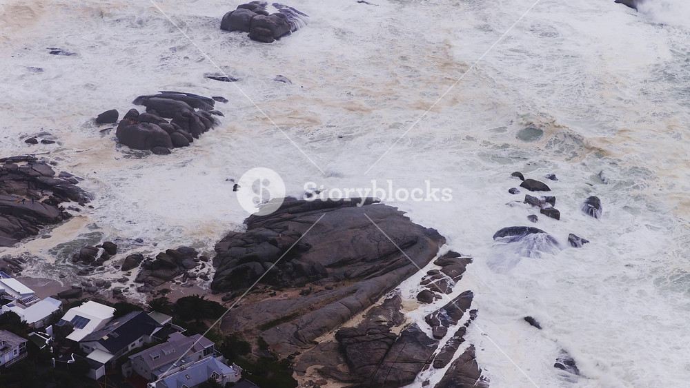 Aerial view of Cape Town coastline with granite boulders and houses. Heavy surf crashes and sweeps white brown foam over rocks in daylight stormy conditions.