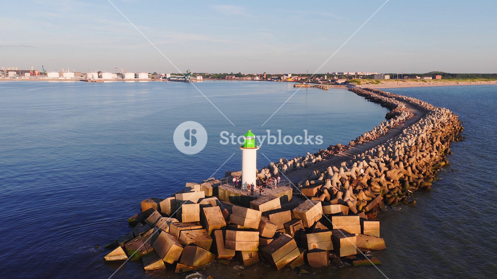 Aerial drone shot shows tetrapod breakwater to white lighthouse with green lantern, people on platform, oil refinery tanks and conveyors, beach and town in Ventspils, Latvia.