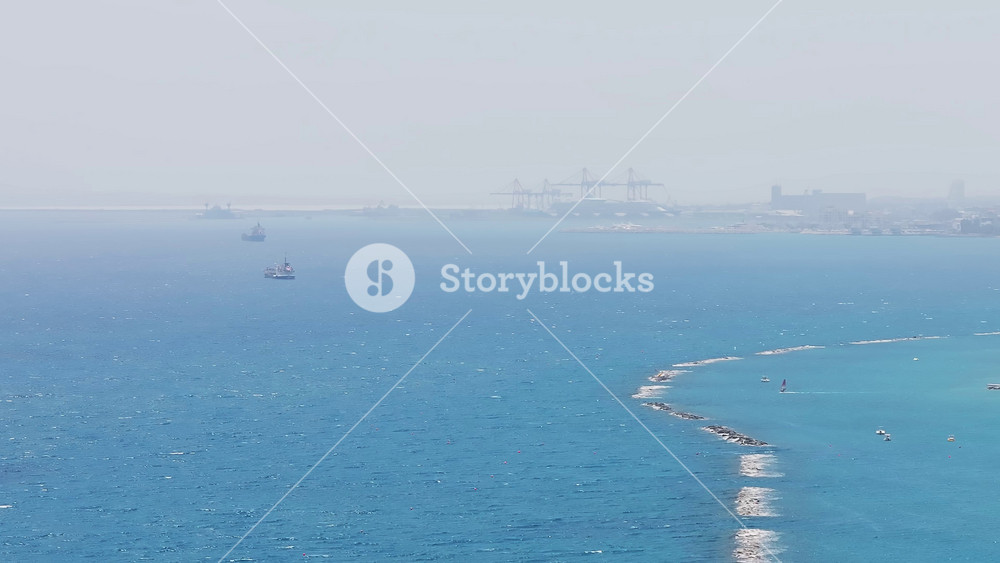 Aerial view of Limassol, Cyprus, showing the coastline, modern buildings, breakwaters, a skyscraper, a port with cranes, and the Mediterranean Sea.