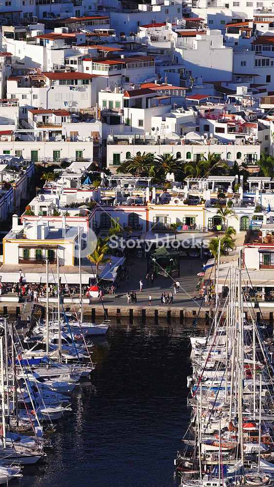 Aerial view shows Puerto de Mogan, Gran Canaria, with SUPERMERCADO VENECIA, palm promenade, restaurants, whitewashed buildings, sailboats, and late afternoon light.