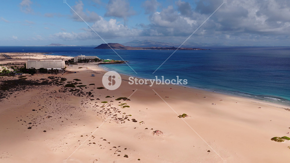 Aerial view of golden dunes and sandy beach near Corralejo, Fuerteventura. Resorts sit by the dunes, Lobos and Lanzarote mark the horizon, waves and clouds move slowly.