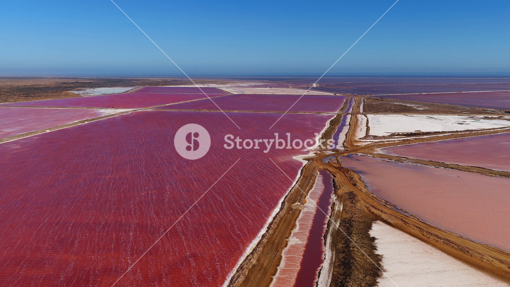 Aerial view of Walvis Bay salt works in Namibia shows red and pink pans, earthen levees, white salt beds, service tracks, salt piles, ocean horizon, and rippling brine.
