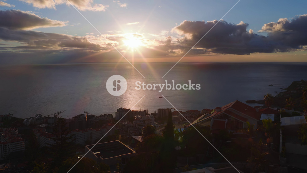 Aerial view of Funchal, Madeira, at sunset. Sunstar reflects on Atlantic water as a sailboat moves. Terraced city, red roofs, pools, cranes, and cliffs appear.