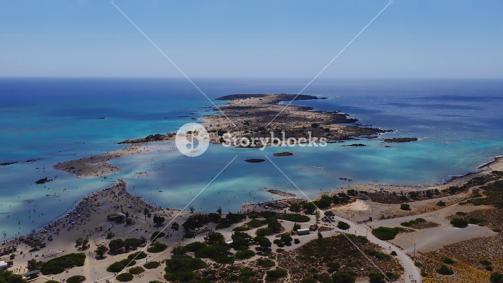 Aerial shot of Elafonisi Beach in Crete, showing pink sand, turquoise waters, shallow lagoons, and visitors wading near a narrow land strip.