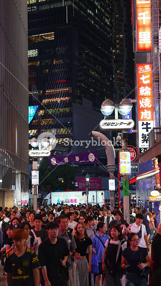 Pedestrians move toward the camera on Shibuya Center gai, Tokyo at night. Neon billboards, a purple banner, and high contrast lighting convey lively nightlife.