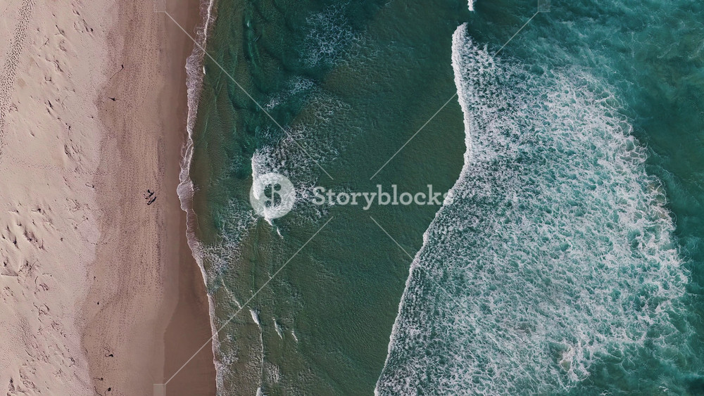Aerial top down view shows pale sand meeting turquoise ocean near Cape Town, South Africa. Waves roll to shore, two figures walk, footprints and sandbars show motion.