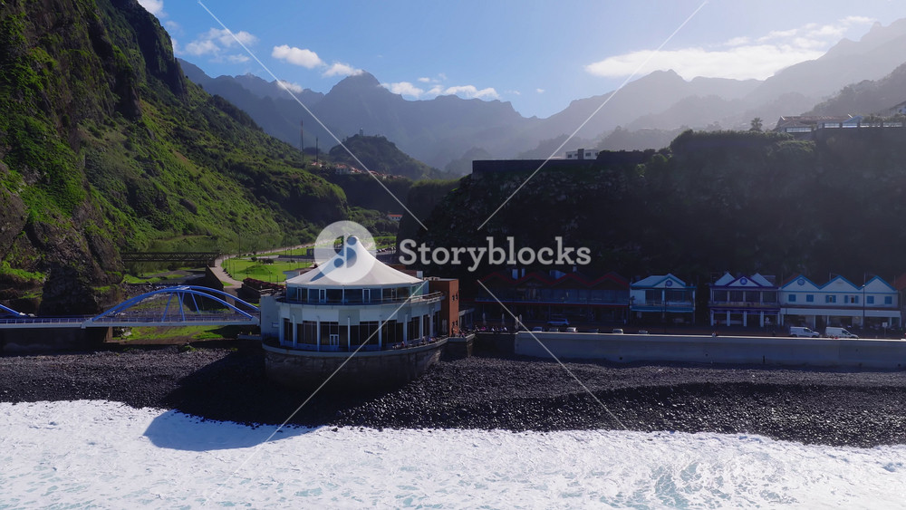 Aerial view shows Madeira coastal village, volcanic pebble beach, white pavilion, blue arched bridge, steep green cliffs, seaside houses, promenade, road, cars, and waves in bright daylight.
