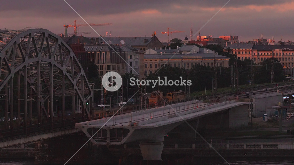 Aerial view of Riga, Latvia shows steel arch railway bridge and new concrete span, cranes, rail tracks, catenary masts, and moving cars at late evening under clouds.