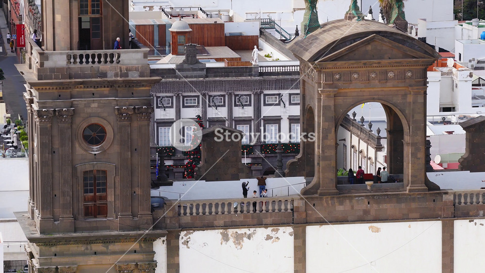 Aerial 4K view of Catedral de Santa Ana, Las Palmas de Gran Canaria, with stone towers, arched loggia, Christmas tree, red stage, flags, palm trees, and people moving.