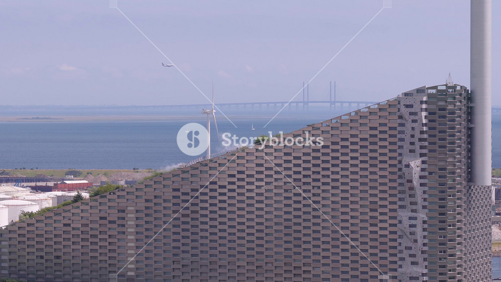 Aerial view of Copenhagen showing Copenhill Power Plant, wind turbines, Oresund Bridge, sailing boats, and rising smoke near the industrial area.