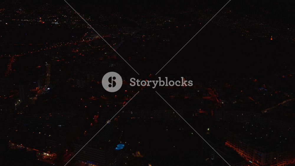 Aerial pan over Funchal, Madeira, Portugal at night shows ridgelines of lights, a purple lit cube atop a modern building, a blue pool, and roads traced by lamps.