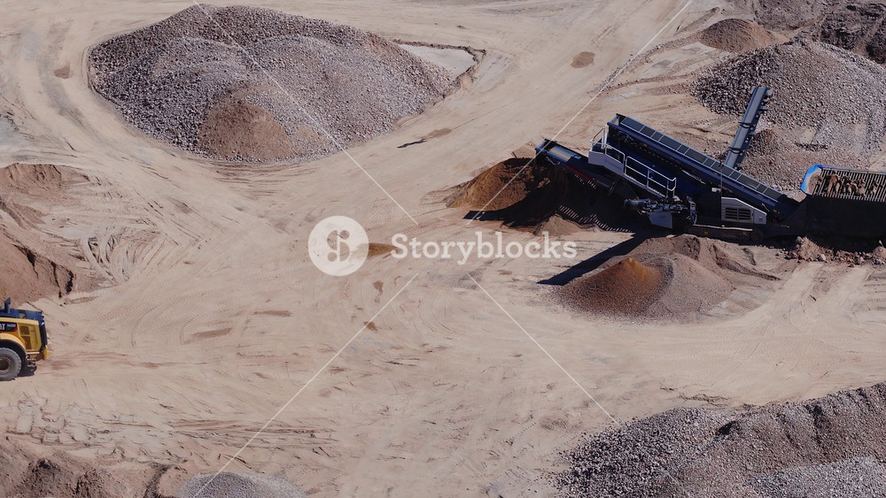 Aerial drone view shows a CAT wheel loader working among gravel and sand piles. Conveyors on a mobile plant form stockpiles as tyre tracks mark the arid ground.