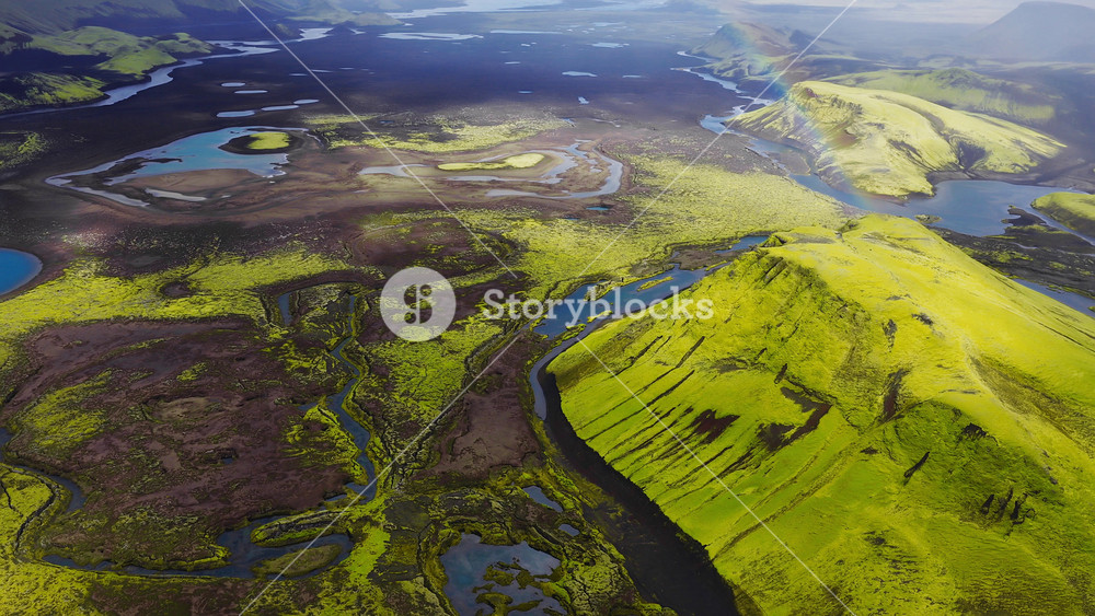 Aerial view in Iceland shows chartreuse slopes, dark lava plains, braided rivers, turquoise lakes, and a faint rainbow under soft daylight near Landmannalaugar.