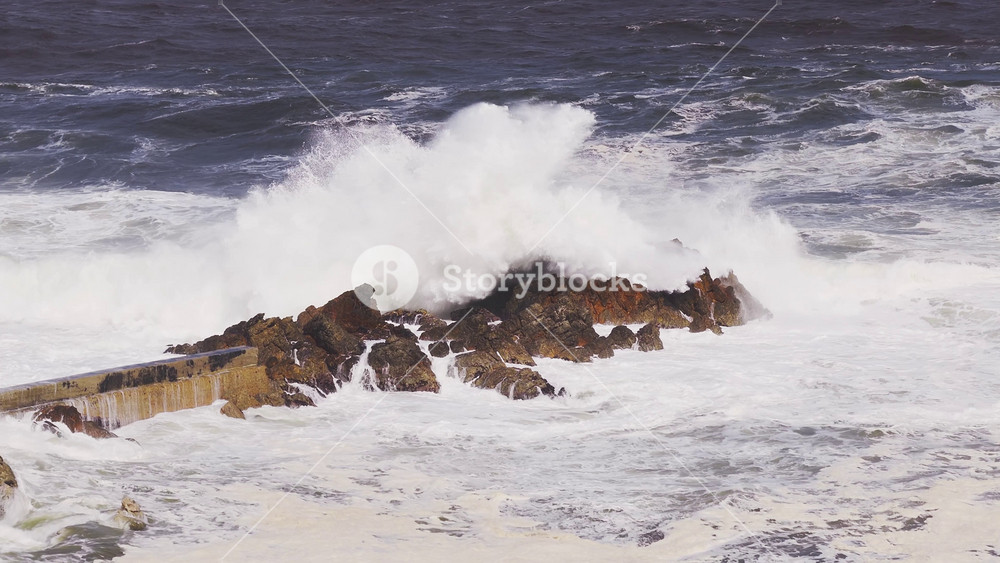 Aerial daytime view of Cape Town, South Africa, near the Cape of Good Hope, as storm swells batter a low concrete breakwater and rocky outcrops with towering white spray.