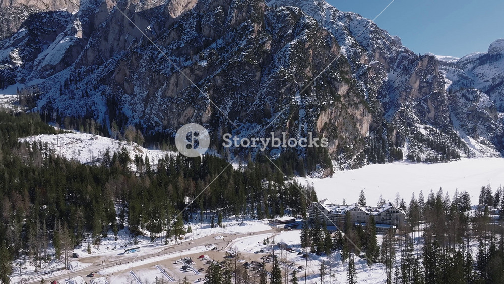 Aerial view of Braies Lake in winter, with a frozen surface, rugged snow dusted mountains, dense evergreen forest, and a nearby lodge with vehicles.