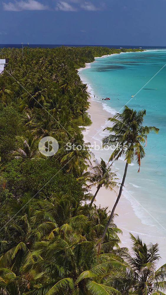 Aerial view of Dhigurah island, Maldives, with a long beach, coconut palms, reef patches, sandbanks, boats, a jet ski, and gentle waves in bright midday light.