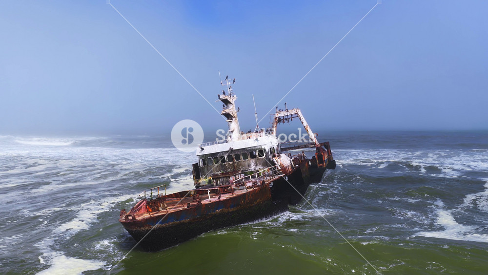 Aerial view shows a rusted Zeila Shipwreck listing in choppy Atlantic surf off Namibia's Skeleton Coast, with seabirds on masts as waves and foam swirl in hazy daylight.