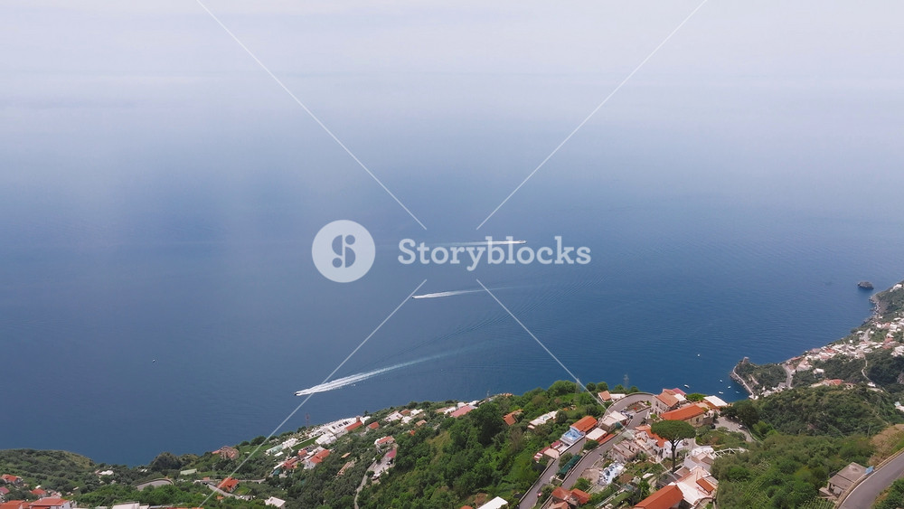 Aerial view of the Amalfi coastline in Italy, showing boats moving on the blue sea and a coastal town with red tiled roofs amid lush greenery.