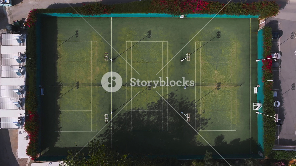 Aerial view of a tennis court complex in Ayia Napa, Cyprus, surrounded by red bougainvillea, parking areas, and pathways, with slow drone motion.