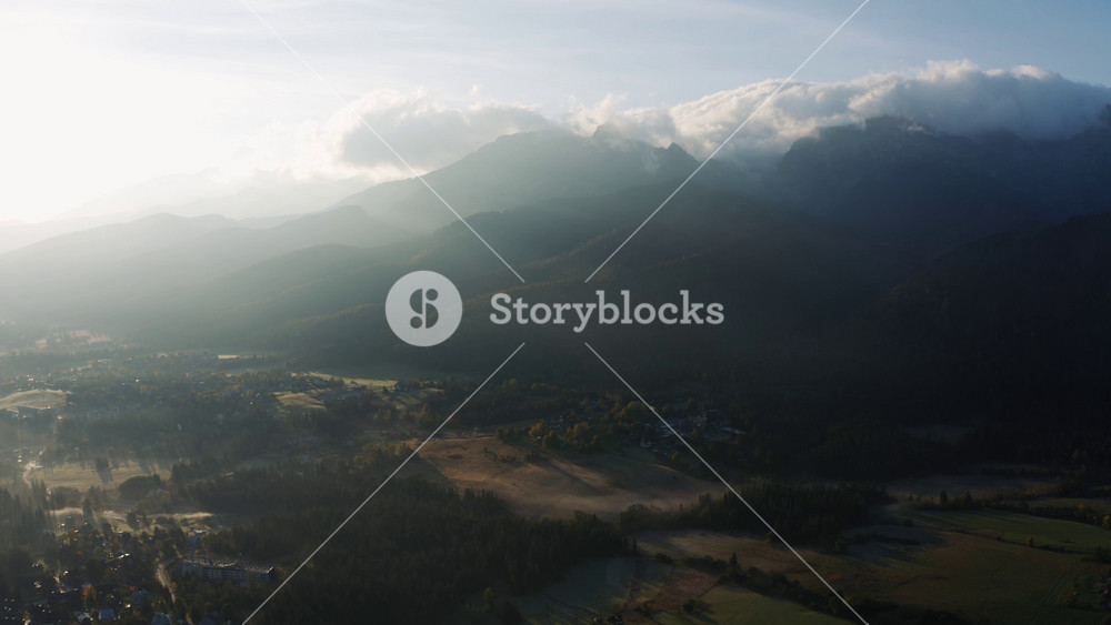 Aerial foggy autumn morning in alpine village lit with bright sunlight. Drone shot epic mountain peaks covered with rolling clouds in sunrise light. Incredible landscape beautiful valley from above