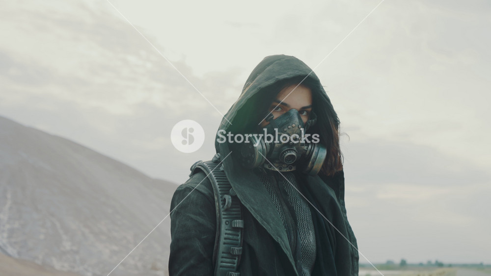 Portrait woman in grunge clothes hooded mantle and protective gas mask posing by white hills looking into camera. Future female survivor. Post apocalyptic world. Steadicam following shot