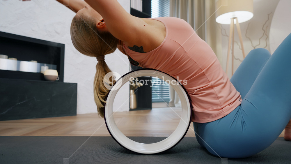 Attractive caucasian woman doing back stretching exercises with yoga wheel on mat in modern living room, female fitness coach practicing yoga training at home, wide angle dolly shot