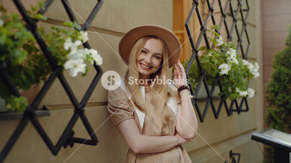 Portrait young cute woman posing outside on sunny evening wearing brown hat leaning against cafe building beige color wall looking at camera smiling. Attractive female relaxing after walk in old town