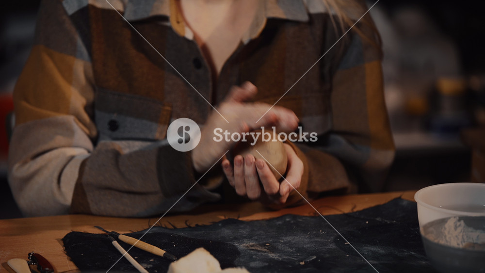 Close-up young female ceramist artist making handmade pottery cup in workshop. Hands of woman potter wearing warm plaid shirt warms up ball piece of clay beats it preparing for modeling
