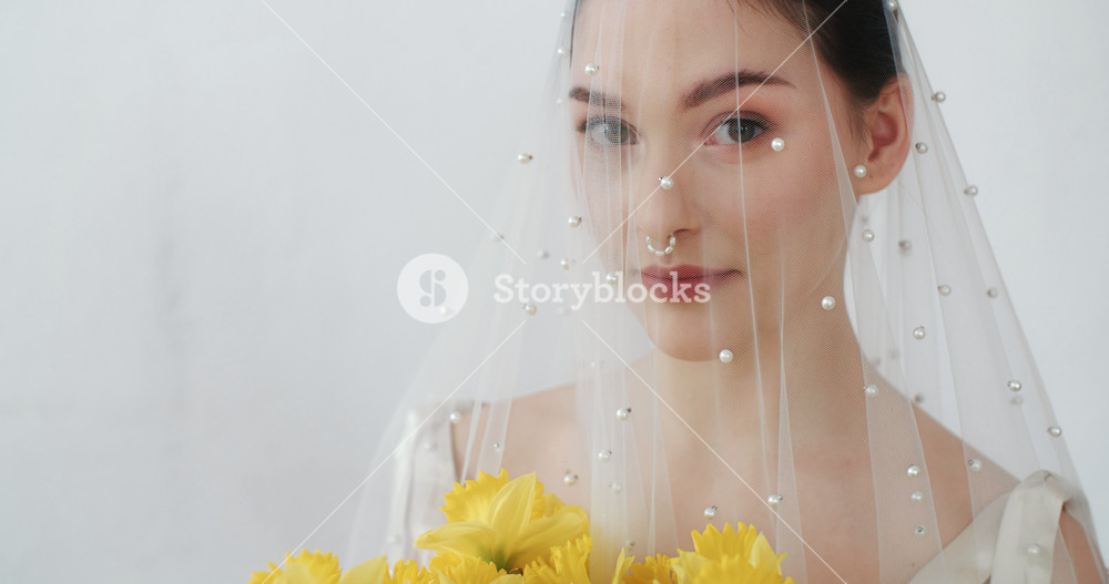 Portrait gorgeous cheerful female holding yellow flowers bouquet looking seductively into camera and smiles standing on clean background. Wedding photo session of sensual bride under white veil