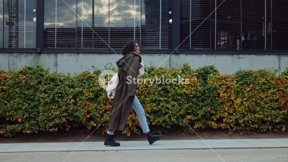 Side view following attractive curly hair woman in autumn outfit with coat on her shoulder walking down and looking around near modern building wall in city street, urban style fashion young female
