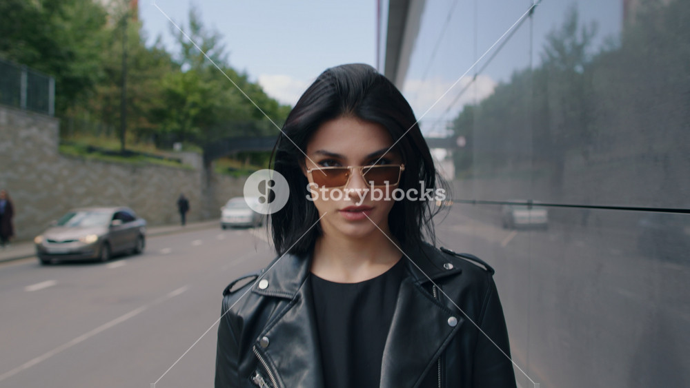 Confident seductive happy brunette female in stylish look black leather jacket and sunglasses walking outdoors along city street looking at camera flirts smiling on sunny day. Urban style fashion