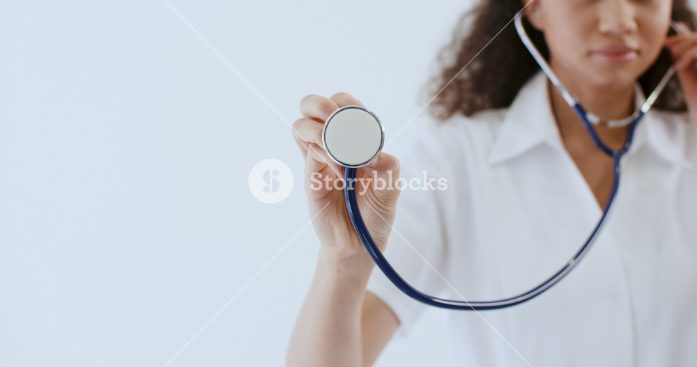 Young African American female doctor in medical coat brings stethoscope to camera lens standing on white background in hospital. Mixed race therapist in uniform. Healthcare and medicine concept