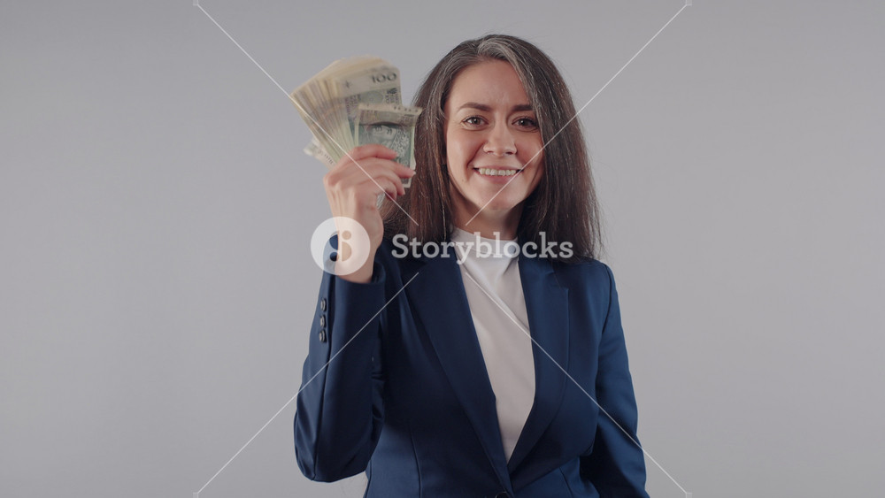 Happy caucasian business woman in blue jacket shows and waving stack of money banknotes bills in her hand standing on grey background, joyful female office manager smiles posing with cash income