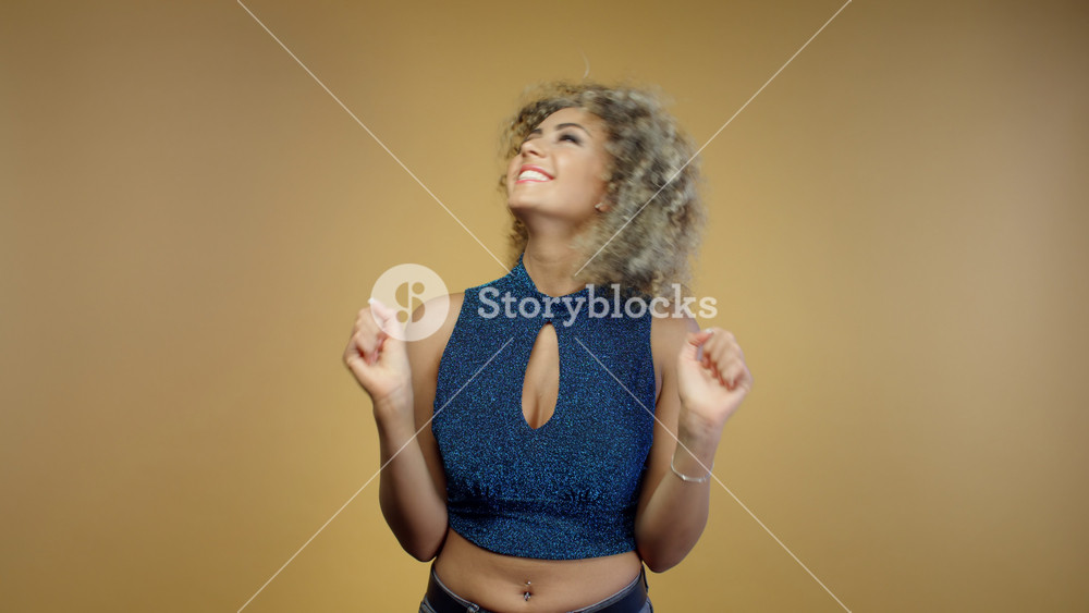 Portrait of joyful young mixed race woman laughing in blue shiny blouse celebrating success on yellow background. Happy hispanic girl jumping and smiling at camera