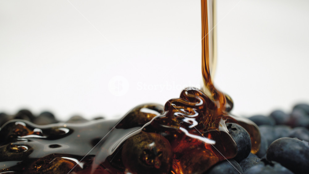 Close-up macro honey pouring on blueberries ripe organic fresh berries on white background dolly slide shot. Caramel flowing, blueberry poured sweet sauce, honey drips over berries